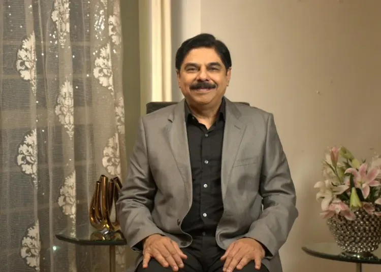 Man in a gray suit sits smiling in a living room, with lace curtains behind and flowers on the side table.